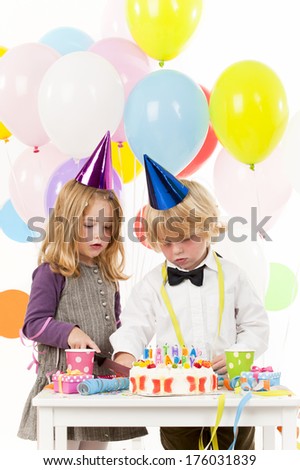 A boy and girl looking at a birthday cake and many balloons over their heads.