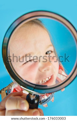A little girl winking into a magnifying glass with one eye closed .