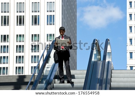A man is standing on an outdoor escalator reading a newspaper.