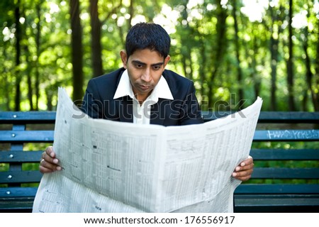 A man in a suit sitting on a bench reading newspaper.