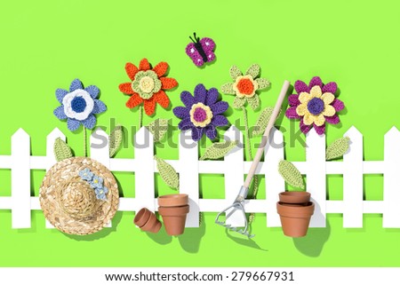 crochet flowers behind a white paper fence with a butterfly, flower pots, rake and a hat on green background