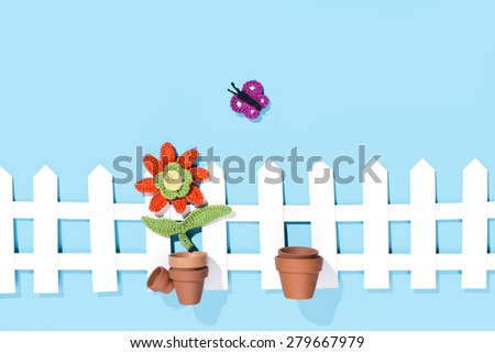 crochet flower with pots, a butterfly and a white fence on blue background