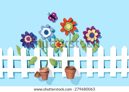 crochet flowers behind a white paper fence with flower pots on blue background