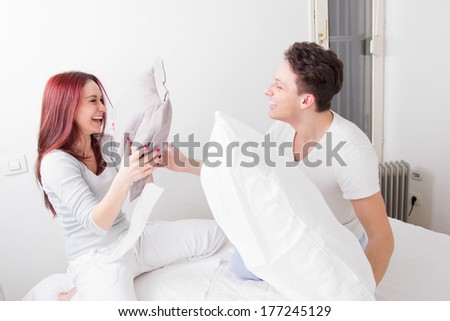 young smiling man and woman fighting with pillows together in bed as sign of happy relationship