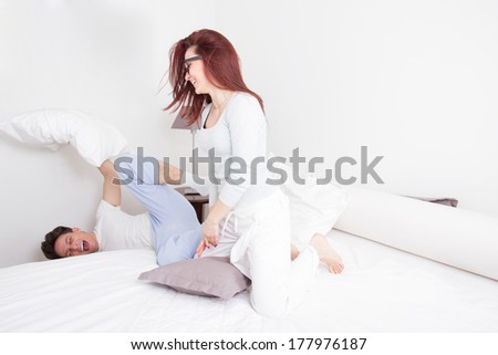 pretty smiling woman in pajamas hitting man with pillow while he falling out of bed