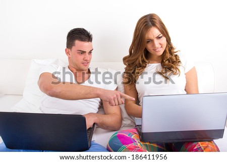 Couple with two laptop computers in bed. Man and woman both with computer, woman upset and surprised man looking at her computer Young modern casual couple in bed. Caucasian man and woman. Funny image