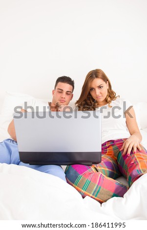 Happy affectionate young married couple sitting side by side in bed relaxing and watching at information on the screen of their laptop. Happy young couple