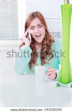 young woman on cellular phone with negative reaction, girl sitting at the table with cup of coffee talking on mobile phone hearing bad news, shocked woman with face expression holding mobile phone