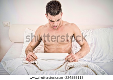 surprised half naked young man in bed  looking down at his underwear at his penis under white covers sheet in badroom. Concept photo of male sexuality and man sex problems, domestic atmosphere.