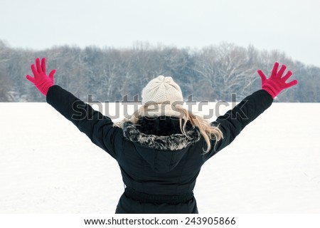 winter woman in snow showing her back and facing forest with hands raised high above, copy space outside on cold winter day.Portrait of happy joyful female model with pink gloves and hat in first snow