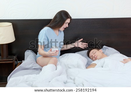 Young woman looking phone while man is sleeping soundly. Jealousy and suspicion in relationship. Caught cheating in messages and calls on mobile in bedroom. Problems in marriage, caucasian couple.
