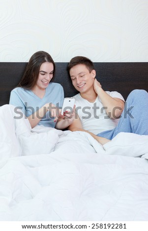 Beautiful young happy couple in bed with mobile phone smiling. Domestic atmosphere, lifestyle photo. Woman shows to man something fanny on mobile