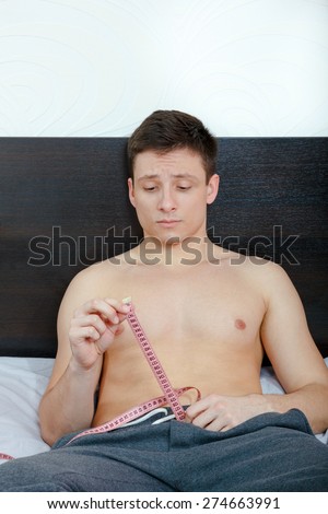 Young handsome half-naked man holding tape measure and measure desired penis length or size while sitting on bed in bedroom in pajamas. Frightened and insecure male model showing length of his pride.