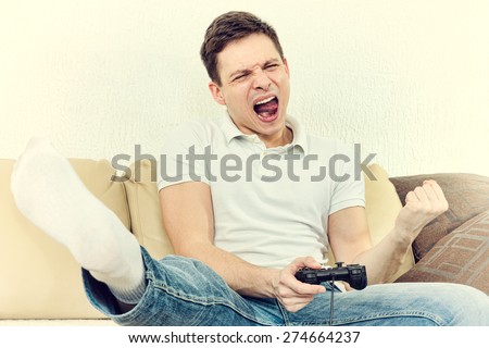 Emotional young man playing video games with joypad or joystick to console or pc. Guy at home in room sitting on bed with thrilled face expression, finally wins his opponent. Photo of winning in game.