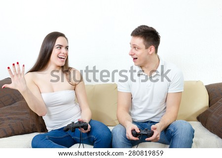Young couple sitting in living room and play video games on console or pc with joysticks while looking in screen or TV. Lifestyle photo of man and woman in jeans at home having fun or entertainment.