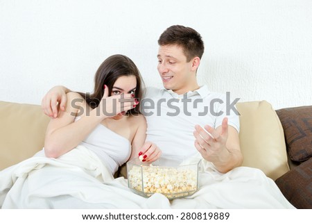 Frightened young attractive couple sitting on the couch in living room at home covered with blanket and watch scary horror on television with popcorn. Terrified woman and smiling man watching movie
