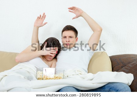 Frightened young attractive couple sitting on the couch in living room at home covered with blanket and watch scary horror on television with popcorn. Terrified man and woman watching violent movie