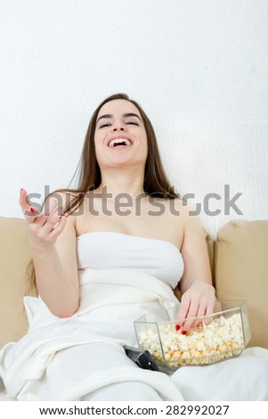 Girl watching movie or TV laughing having fun eating popcorn in bed. Alone young Caucasian woman model sitting down on bed.  Smiling female watching funny comedy in living room at home.