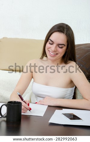 Leasure and home concept - calm teenage girl woman writes with pen on white paper and sitting on couch at home. Smiling female model writing something on table and sitting on bed at living room.