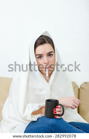 Upset woman with thermometer in her mouth sick colds, flu, fever and migraine in bed with black cup of tea or coffee. Girl with virus. Female model caught cold covered with white blanket at home.
