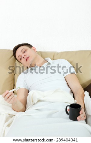 Sick man with fever laying in bed measuring temperature with thermometer and holding cup of tea