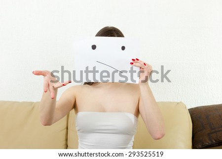 Young woman is confused. Girl doesn\'t know what to do. Girl holds forefinger and smiley drown on paper. Young  female bears sad face drawing on white sheet. Wonering about things.