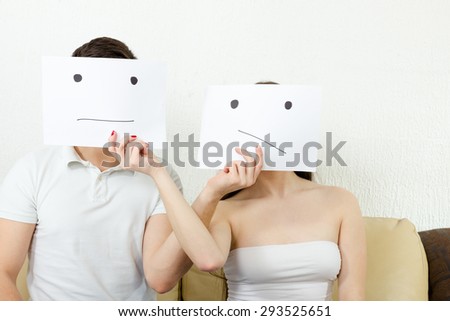 Worrying  upset young couple struggle with troubles. Concerned teenagers in despair. Unhappy persons sitting on couch. Man and woman cover their faces with sad smiley  drawn on paper with one tear.