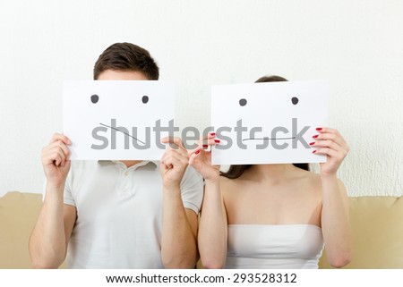 Worrying young couple  is very concerned and having troubles. Teenagers in despair. Unhappy persons sitting on couch. Man and woman cover their faces with sad smiley  drawn on paper with one tear.