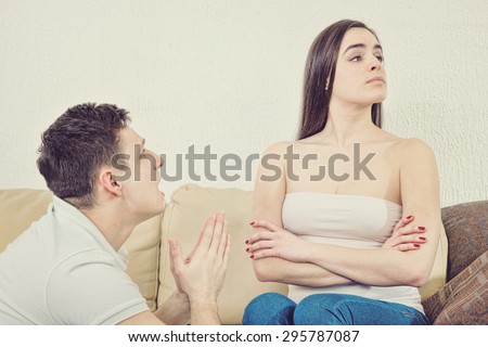 Portrait of couple, man and woman. Sad husband on his knees asking, begging for forgiveness wife who refuses to accept apology on sofa at home. Human emotions, face expressions.