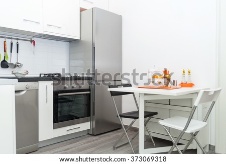 Interior of Small White Kitchen with Fresh Fruit Basket on White Table With Two Chairs. Bright Modern Kitchen Interior Background. Must Have Kitchenware and Appliances, Stainless Fridge, Stove, Sink.