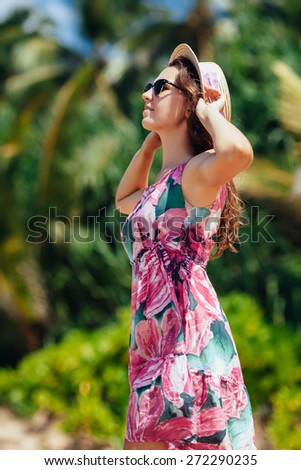 Young woman standing on a beach holding her straw hat and looking to the horizon.Palm background