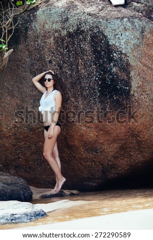 Young girl in black swimsuit and white  blouse on the rocks near the sea beach.full-length portrait