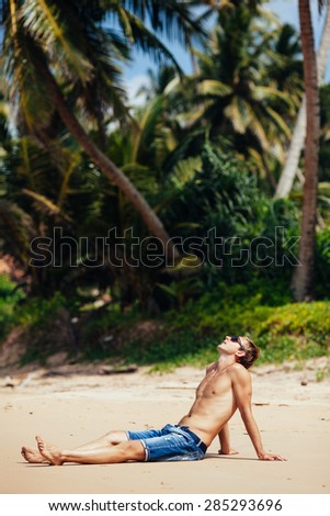 Man relaxing on a tropical beach. Young tanned man taking sunbath on sand beach with many palm trees