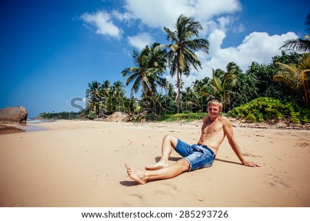 Man relaxing on a tropical beach. Young tanned man taking sunbath on sand beach with many palm trees