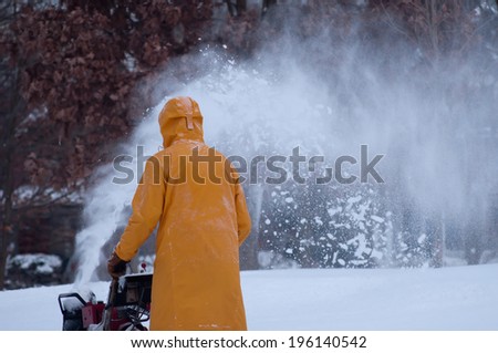 A man in a yellow snow coat uses his red snowblower to clear his sidewalk of snow.