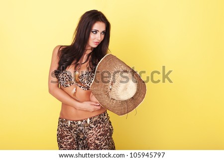 Fashionable brunette in two piece summer leopard print outfit holding a large wide brimmed straw hat against a yellow studio background