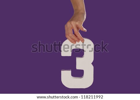 Female hand holding up the number 3 against a purple background conceptual of numbers, measurement, amount, quantity, accounting and mathematics