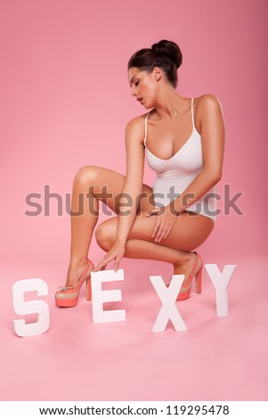 Sensual sexy woman in a swimsuit posing crouching on the floor in stilettoes with the word SEXY in white capital letters in front of her on a pink background
