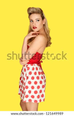 Glamorous blonde retro fashion model in a red polka dot miniskirt with vivid red lipstick and an olden day coiffure posing against a yellow studio background