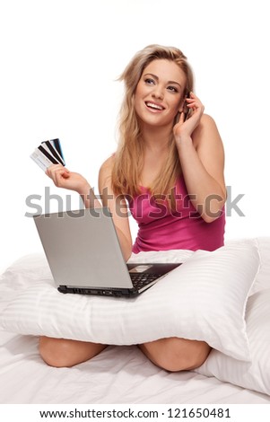 Happy online shopper chatting on her mobile while sitting with her laptop balanced on a pillow on her lap and a fistful of credit cards