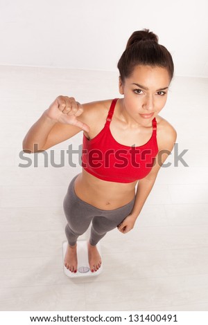 High angle view of an athletic young woman in sportswear standing on a scale giving a thumbs down gesture of disappointment and disapproval