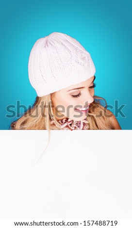 Beautiful young woman wearing a white knitted cap holding a blank white sign and looking down at it with a delightful smile