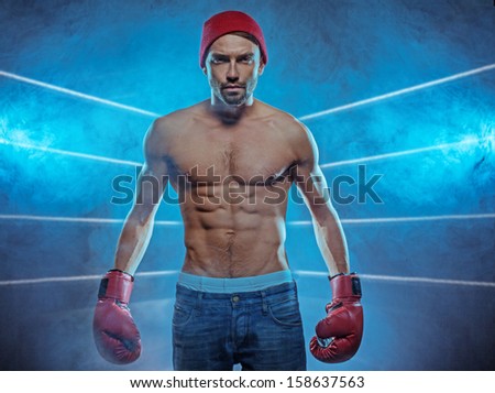 Young athletic boxer exhibiting his six-pack abdominals wearing gloves, blue jeans and a beanie hat in a smoky background