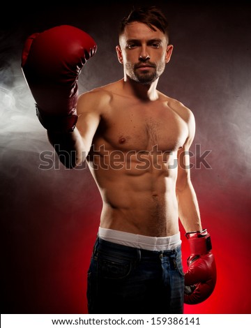 Young bearded male boxer with a muscular physique holding up his fist at the camera in a red leather boxing glove on a shadowed red studio background