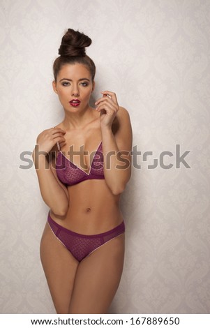 Beautiful busty young woman in sexy trendy purple lingerie with her hair in a neat bun standing looking at the camera with a seductive sultry expression