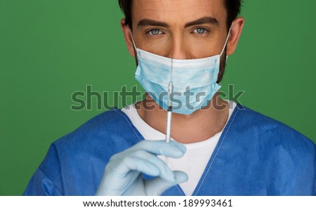Doctor or male nurse wearing a surgical mask holding up his hand with a hypodermic syringe ready to give a patient an injection, close up view on green with copyspace