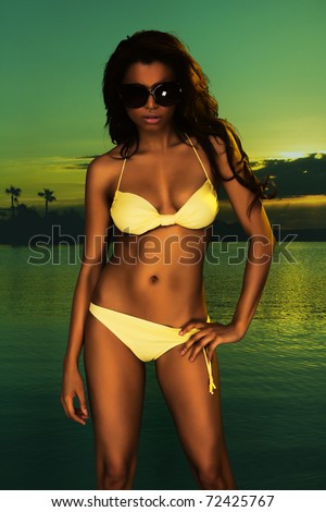 Beautiful young brunette model wearing yellow bikini photographed against colorful sunset over the beach - studio