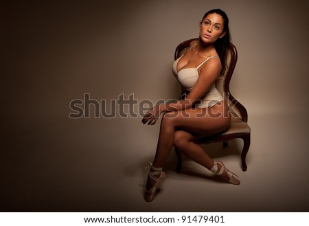 Dramatic Portrait Of Sexy Ballerina sitting sideways on a vintage chair facing towards camera and displaying large breasts and cleavage, dark background with ballerina highlighted,