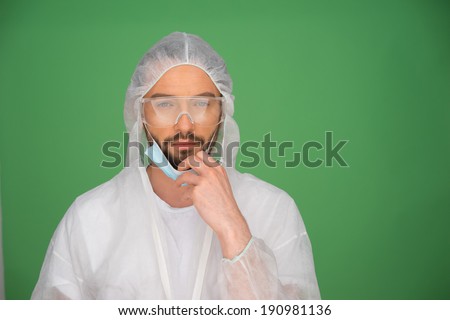 Bearded young male laboratory technician in a sterility suit, goggles and a mask on a green background with copyspace