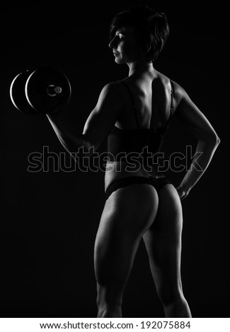 Dark portrait of a beautiful muscular sexy female body builder standing sideways in lingerie displaying her curvaceous buttocks while lifting weights